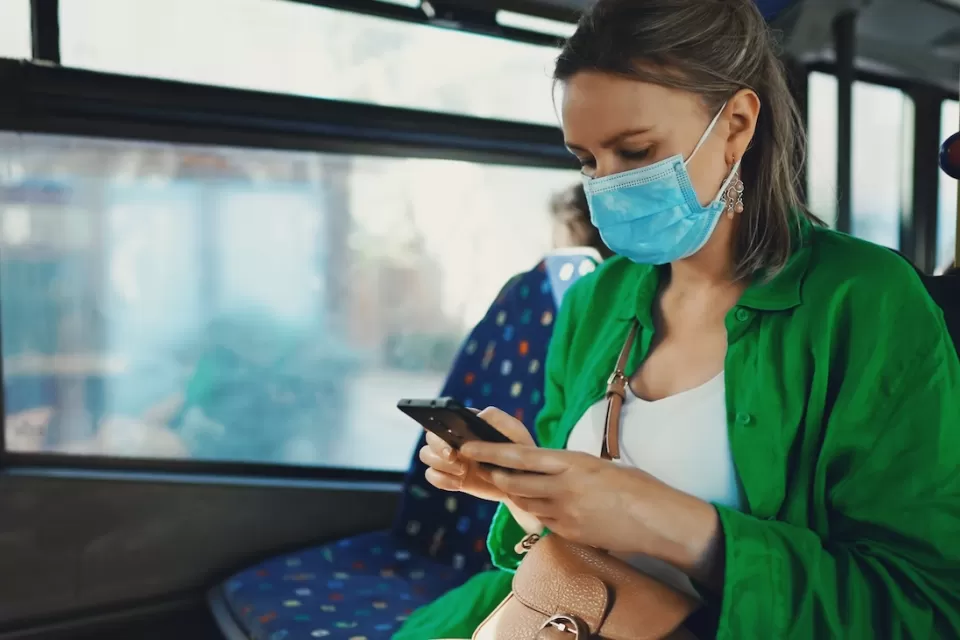 Woman in green sweater wearing light blue medical mask sitting on bus looking at her phone.